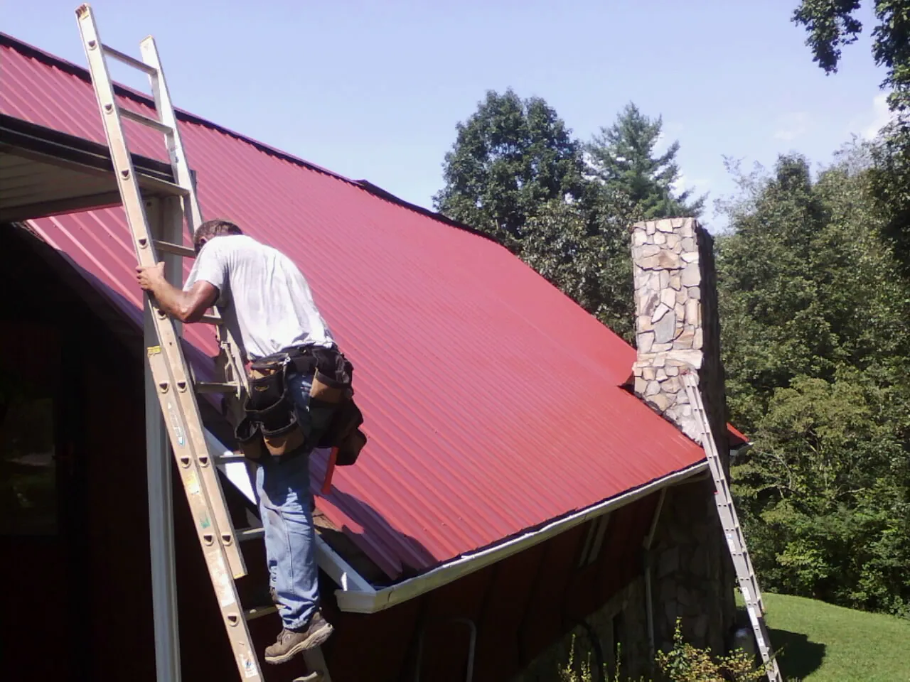 Roofer on a ladder beside a red metal roof and stone chimney — Soffit & Fascia Repair by Roofing Brothers in Clarkfield